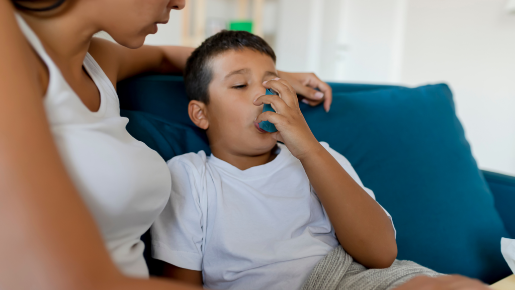 boy using an inhaler to control asthma and wheezing sitting on a couch with his mother